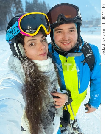 Couple enjoying a winter day on the ski slopes in a snowy mountain landscape with ski gear and equipment amidst beautiful scenery Couple enjoying a winter day on the ski slopes in a snowy mountain landscape with ski gear and equipment amidst beautiful scenery 133044116
