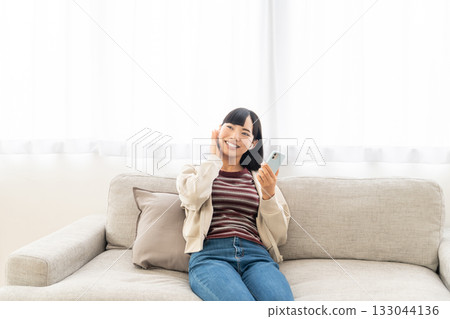 年輕女子坐在沙發上,用智慧型手機聽音樂。 年輕女子坐在沙發上,用智慧型手機聽音樂。 133044136