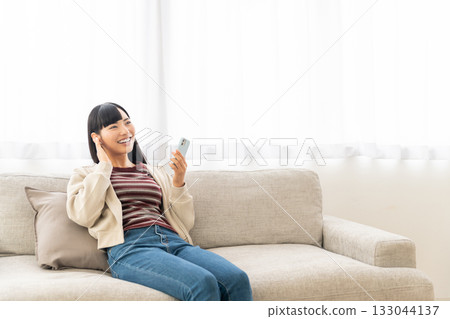 年輕女子坐在沙發上，用智慧型手機聽音樂。 133044137