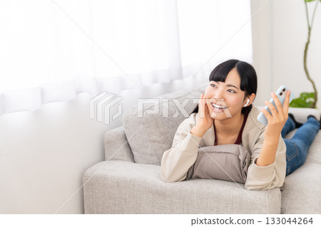 A young woman lying on a sofa and listening to music on her smartphone A young woman lying on a sofa and listening to music on her smartphone 133044264