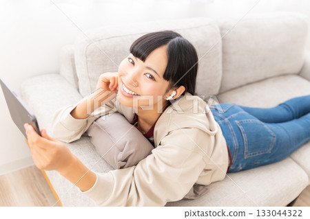 A young woman lying on a sofa watching a video on an iPad 133044322