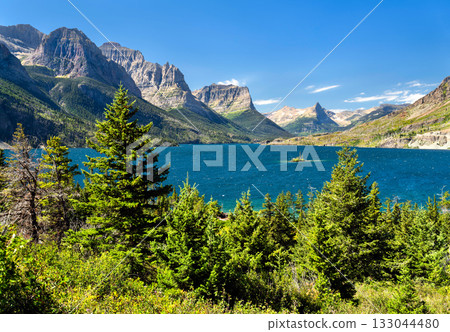Panoramic view of the vibrant blue Saint Mary Lake, featuring Wild Goose Island, in Glacier National Park. The Rocky Mountains rise grandly under a clear blue sky, framed by pine trees 133044480