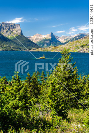 Vertical view of Wild Goose Island in the deep blue Saint Mary Lake, Glacier National Park. The Rocky Mountains rise in the background, framed by pine trees under a blue sky Vertical view of Wild Goose Island in the deep blue Saint Mary Lake, Glacier National Park. The Rocky Mountains rise in the background, framed by pine trees under a blue sky 133044481
