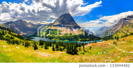 A wide panoramic view of Hidden Lake and Bearhat Mountain from the Logan Pass overlook in Glacier National Park, Montana. A green meadow is in the foreground under a dramatic cloudy sky 133044482