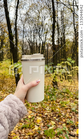 Hand holding a beige thermal mug in a beautiful autumn forest with yellow leaves. Warm fall mood Hand holding a beige thermal mug in a beautiful autumn forest with yellow leaves. Warm fall mood 133044642