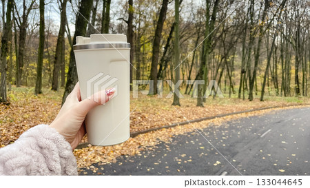 Hand holding a thermal mug on an autumn walk. Horizontal shot of a park path with fallen leaves Hand holding a thermal mug on an autumn walk. Horizontal shot of a park path with fallen leaves 133044645