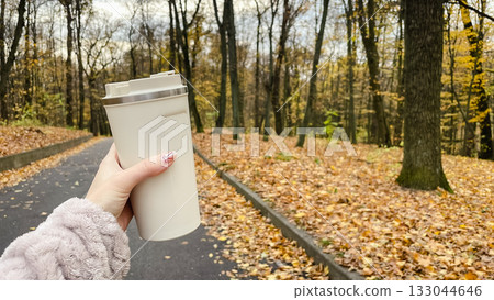 Hand holding a thermal mug on an autumn walk. Horizontal shot of a park path with fallen leaves Hand holding a thermal mug on an autumn walk. Horizontal shot of a park path with fallen leaves 133044646