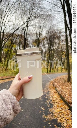 Woman's hand holding a beige travel mug while walking on an autumn park path covered with leaves Woman's hand holding a beige travel mug while walking on an autumn park path covered with leaves 133044654