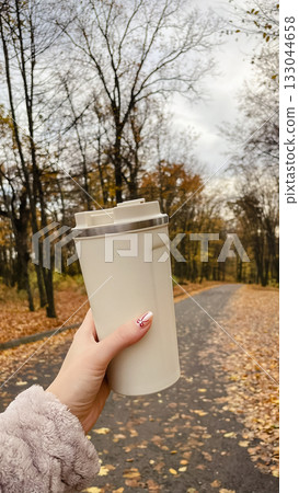 Woman's hand holding a beige travel mug while walking on an autumn park path covered with leaves Woman's hand holding a beige travel mug while walking on an autumn park path covered with leaves 133044658