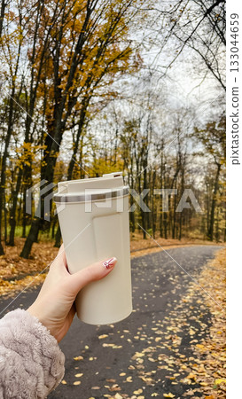 Woman's hand holding a beige travel mug while walking on an autumn park path covered with leaves 133044659