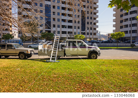 Yerevan Armenia 10.22.2025. A white truck with a ladder on top of it is parked in a grassy area next to a bu 133046068