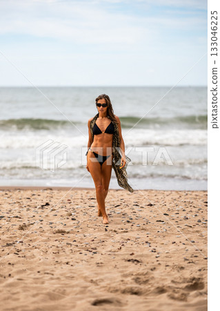 A stylish model gracefully strutting along the soft sandy beach, wearing a chic black bikini and trendy sunglasses 133046225