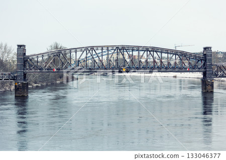 Snowy morning at Magdeburg Schleinufer shows calm river Elbe reflecting steel Hubbrucke bridge. Quiet winter path lined with blue lamps and benches creates serene urban landscape 133046377
