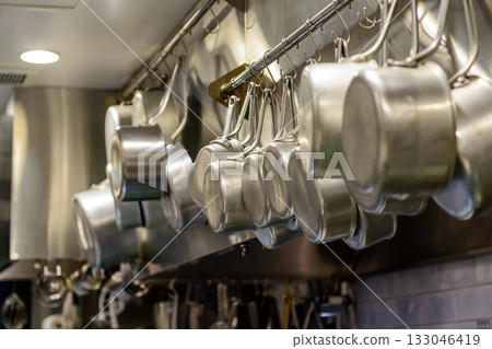 Stainless steel pots lined up in a restaurant kitchen, cooking utensils hung in an orderly fashion Stainless steel pots lined up in a restaurant kitchen, cooking utensils hung in an orderly fashion 133046419