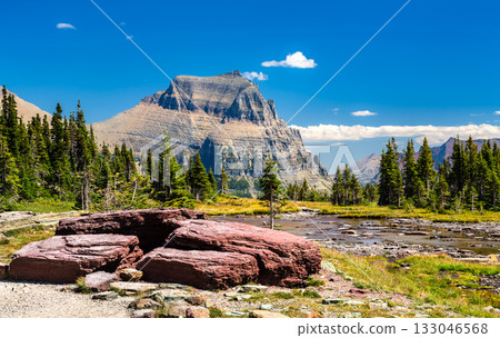 Scenic view of Mount Oberlin from Logan Pass in Glacier National Park. Red argillite rocks are in the foreground, next to an alpine stream and pine trees, under a clear blue sky Scenic view of Mount Oberlin from Logan Pass in Glacier National Park. Red argillite rocks are in the foreground, next to an alpine stream and pine trees, under a clear blue sky 133046568