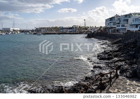 A scenic view of a coastal town in Lanzarote, showcasing the marina, boats, and the rugged shoreline under a cloudy sky. A scenic view of a coastal town in Lanzarote, showcasing the marina, boats, and the rugged shoreline under a cloudy sky. 133046982