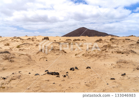 A scenic view of a volcanic mountain on La Graciosa, with sandy dunes and sparse vegetation under a bright cloudy sky. 133046983