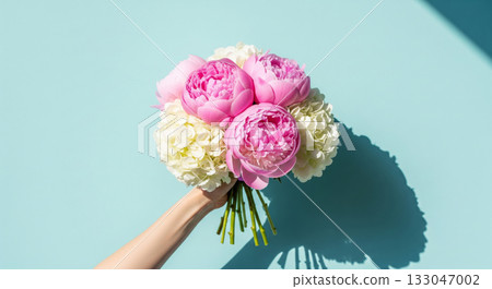 A hand holds a beautiful, elegant bouquet of fresh flowers against a bright, light blue background 133047002