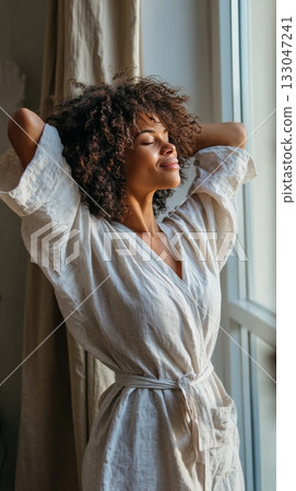 Person in a linen robe enjoys a peaceful moment by the window, stretching and embracing self-care in a calming home environment 133047241