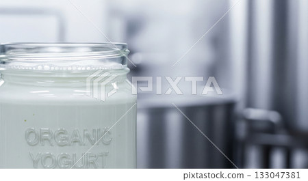 Organic Greek yogurt sits smoothly in a clear glass jar, showcasing its creamy texture. A steel kitchen backdrop highlights this fresh dairy product, emphasizing its homemade appeal. 133047381