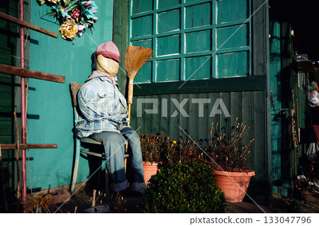 A scarecrow sitting on a wooden chair in front of a small garden gazebo on an allotment, cleverly imitating a resting human figure, basking peacefully in the warm sunlight of a golden autumn day. 133047796