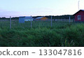 A farm plot with outbuildings and residential buildings behind a chain-link fence with an unmown lawn at dusk, a red farmhouse and barns on a private farmland, on a windy summer evening without people 133047816