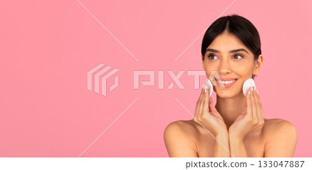 Smiling woman cleaning face with cotton pads on pink background Smiling woman cleaning face with cotton pads on pink background 133047887