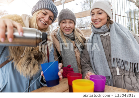 Portrait three pretty young adult woman friends enjoy laughing pour hot mulled wine thermos at Christmas market outdoors cold winter day. People having fun celebrate new year time street cafe outside Portrait three pretty young adult woman friends enjoy laughing pour hot mulled wine thermos at Christmas market outdoors cold winter day. People having fun celebrate new year time street cafe outside 133048012