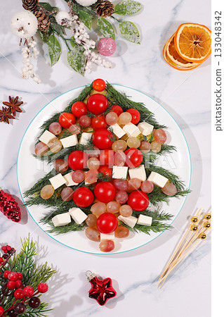 Traditional holiday salad in the form of a live edible Christmas tree with feta cheese, olives, tomatoes and grapes, Christmas and New Year's dishes, food design idea, fir branches and decorations 133048342