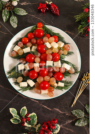 Christmas and New Year's dishes, traditional holiday salad in the form of a live edible Christmas tree with feta cheese, olives, tomatoes and grapes, fir branches and decorations, food design idea Christmas and New Year's dishes, traditional holiday salad in the form of a live edible Christmas tree with feta cheese, olives, tomatoes and grapes, fir branches and decorations, food design idea 133048346