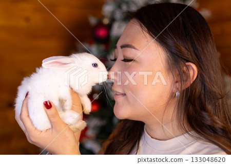 A woman kisses a white rabbit in front of a Christmas tree, A woman kisses a white rabbit in front of a Christmas tree, 133048620
