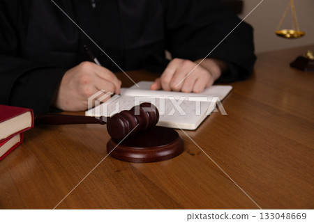 A judge writing notes on a book next to a gavel on a wooden desk in a courtroom  133048669