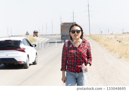 A woman walks along a road next to a white car 133048695
