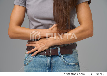Close-up of woman s hands clutching stomach in pain, studio shot on neutral grey Close-up of woman s hands clutching stomach in pain, studio shot on neutral grey 133048702