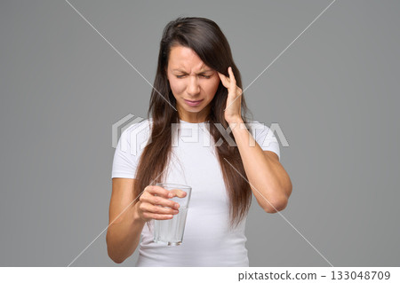 Woman with headache holding glass of water expressing pain or discomfort against gray background 133048709