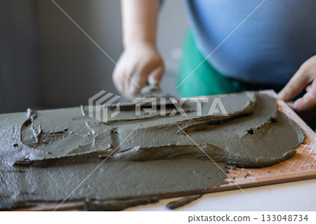 A worker applies cement tile adhesive to the back of a tile with a trowel, preparing it for installation during a home renovation or construction project. Applying adhesive for tile installation. A worker applies cement tile adhesive to the back of a tile with a trowel, preparing it for installation during a home renovation or construction project. Applying adhesive for tile installation. 133048734