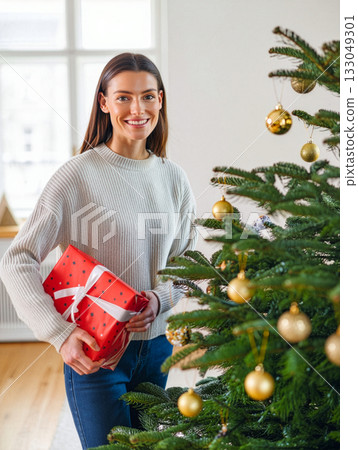 Smiling woman holding a red gift box near a decorated Christmas tree in a cozy living room during the holiday season 133049301