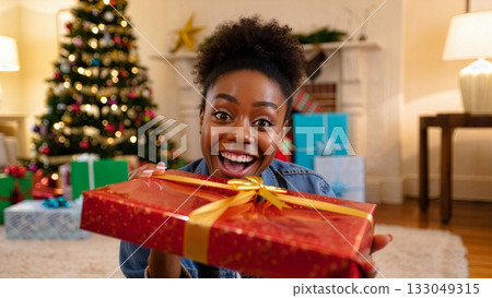 Excited person holding a red gift with a golden ribbon in front of a decorated Christmas tree during holiday celebrations at home Excited person holding a red gift with a golden ribbon in front of a decorated Christmas tree during holiday celebrations at home 133049315
