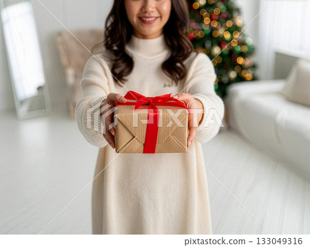 Gift giving moment captured during a cozy indoor holiday setting with a smiling woman in a soft sweater and a beautifully decorated Christmas tree in the background 133049316