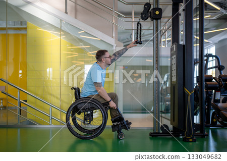Man in wheelchair using fitness equipment for arms strength buildup at post injury session in gym. Man in wheelchair using fitness equipment for arms strength buildup at post injury session in gym. 133049682