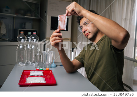 Skilled agronomist wearing safety glasses examining fertilizer packet while preparing to nourish plants in vertical hydroponic tower, surrounded by measuring equipment and recycled plastic bottles. 133049756
