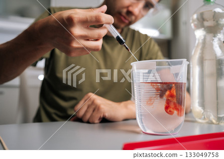 Cropped shot of agronomist male in safety glasses adding red liquid fertilizer to water in beaker using syringe, preparing solution for plant growth and nutrition. Concept of home gardening. 133049758
