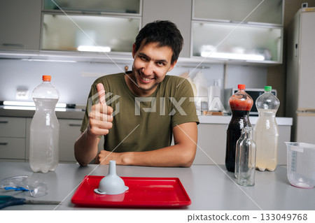 Cheerful gardener giving thumbs-up gesture to indicate success or approval sitting at table in kitchen, preparing fertilizer for plants using various liquids and funnel, smiling looking at camera. Cheerful gardener giving thumbs-up gesture to indicate success or approval sitting at table in kitchen, preparing fertilizer for plants using various liquids and funnel, smiling looking at camera. 133049768