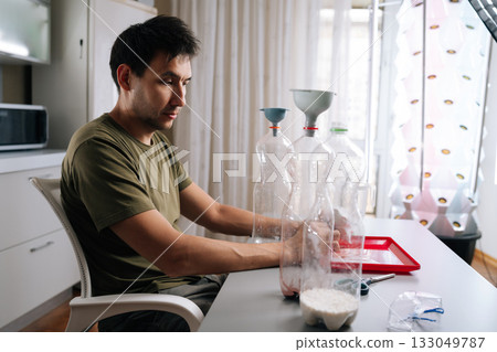 Side view of gardener mixing fertilizer for plants in vertical hydroponic tower garden, using recycled plastic bottles and funnels in home environment. Concept of healthy and sustainable cultivation. 133049787
