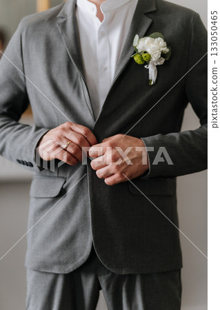 Groom buttoning grey suit with boutonniere, wedding ring closeup 133050465