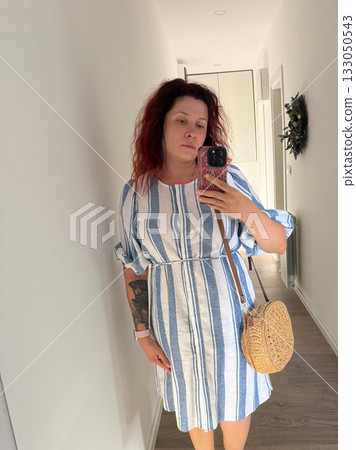 Woman taking mirror selfie in striped linen dress with straw bag. Minimal style, everyday authenticity, and modern self-expression through fashion. 133050543
