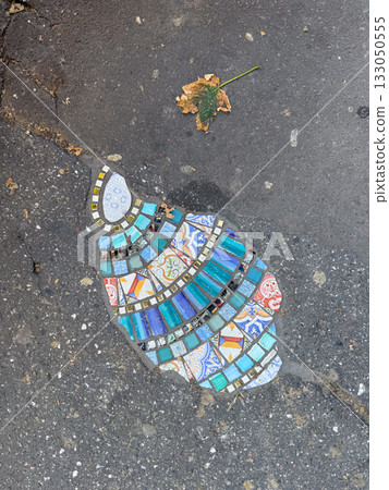 Colorful street mosaic embedded in asphalt with fallen leaf nearby. Urban art, creativity, and poetic contrast between decay and expression. 133050555