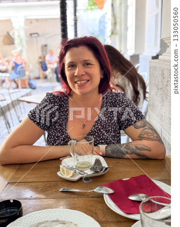 Woman sitting at wooden table in cozy outdoor cafe, smiling in warm sunlight. Relaxed atmosphere, everyday joy, and friendly human connection. 133050560