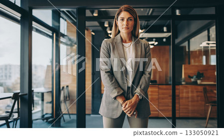 A business woman in a smart outfit stands in a modern office with large windows. She looks ready for a meeting, showcasing professionalism and confidence in a stylish workspace. 133050921