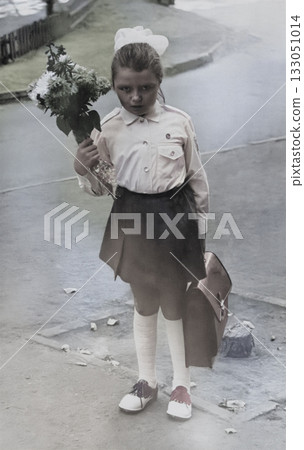 A retro portrait of a second-grade girl in a formal school uniform holding a bouquet of flowers and a school bag. Vintage photo from 1975. A retro portrait of a second-grade girl in a formal school uniform holding a bouquet of flowers and a school bag. Vintage photo from 1975. 133051014